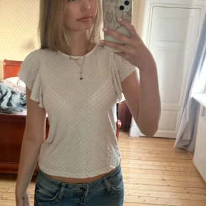 Vit prickig blus med volangärmar - Supersöt vit blus med små beige/guldiga prickar och volangiga korta ärmar. Toppen har rund halsringning och är gjord i mjuk bomull, perfekt för en fräsch och luftig look. Passar till jeans eller kjol för en chill stil.