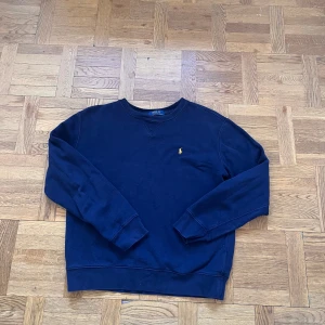 Mörkblå sweatshirt från Polo Ralph Lauren - Klassisk mörkblå sweatshirt från Polo Ralph Lauren med rund halsringning och broderad gul logga på bröstet. Tröjan har långa ärmar och ribbade muddar vid ärmslut och nederkant. Perfekt för en chill och stilren look.