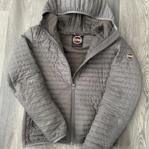 Grå pufferjacka från Colmar, strl 52 - Snygg grå pufferjacka från Colmar med huva och quiltad design. Materialet är polyester och jackan har långärmad passform. Perfekt för kyliga dagar. Några defekter se bild 4 och 5!