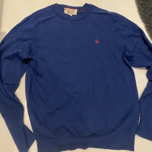 Blå tröja från Penguin Munsingwear - Snygg blå tröja från Penguin by Munsingwear, tillverkad i Italien. Tröjan har ett litet rött pingvinbroderi på bröstet och rund hals. Materialet är mjukt och lätt, perfekt för dig som gillar klassisk stil med en twist. Långärmad och med ribbade muddar. Tröjan står att det ska vara en L men den är liten i storleken så den passar M