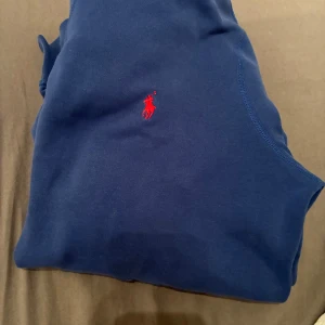 Blå hoodie från Polo Ralph Lauren - Snygg marinblå hoodie från Polo Ralph Lauren med klassisk röd broderad logga på bröstet. Tröjan har huva med vit snörning och en stor magficka. Tillverkad i mjuk bomull, perfekt för chill dagar eller när du vill ha en clean streetstyle.