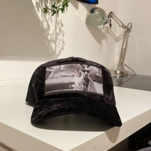 Svart keps från Hatstore med tryck - Svart truckerkeps från Hatstore med mesh på sidorna och justerbar snapback. Framtill finns ett svartvitt tryck med motiv av en kengru i solglasögon och jacka framför en bil. Kepsen har böjd skärm och cool streetstil.