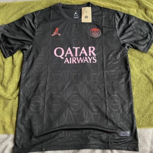 PSG x Jordan svart fotbollströja - Snygg svart PSG fotbollströja i samarbete med Jordan. Tröjan har diskret mönster, rosa logga och text, samt Paris Saint-Germain-märke på bröstet. Tillverkad i lätt och ventilerande Dri-FIT-material som passar perfekt till träning eller match.