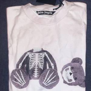 Palm Angels vit t-shirt med björntryck - Palm Angels t-shirt i vit bomull med stort tryck av en björn med skelettdetaljer och avskild huvuddel på framsidan. Klassisk rund hals och korta ärmar. Palm Angels-logga under motivet. Perfekt statement-plagg för dig som gillar streetwear och edgy design. Helt ny oanvänd om du är intresserad lägg ett prisförslag eller om du har några frågor så kan du fråga.