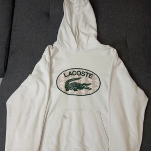 Vit hoodie från Lacoste med logga - Snygg vit hoodie från Lacoste med stor grön krokodil-logga och text på bröstet. Klassisk känguruficka framtill och huva. Perfekt för dig som gillar streetwear och vill ha en clean look med ikonisk branding.