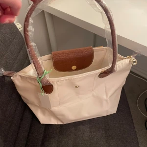 Beige axelväska från Longchamp - Snygg beige axelväska från Longchamp med bruna skinnhandtag och lock. Väskan har guldfärgade detaljer och dragkedja upptill. Perfekt storlek för att få plats med allt du behöver och klassisk design som alltid funkar. Original priset på väskan är 800 kr.