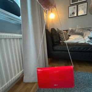 Röd kuvertväska från DKNY med kedja - Snygg röd kuvertväska från DKNY i glansigt skinn med silverfärgad kedja som axelrem. Väskan har flera kortfack  på insidan, perfekt för att hålla ordning på allt viktigt. Stilren och modern design med DKNY-logga framtill.