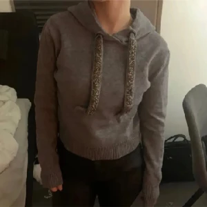 Zara Grå croppad hoodie med glitterband - Zara Grå croppad hoodie med huva och långa ärmar. Tröjan har breda snören med glitterdetaljer som ger en extra touch. Ribbad kant vid midjan och ärmslut. Perfekt för dig som gillar chill och snygg stil.