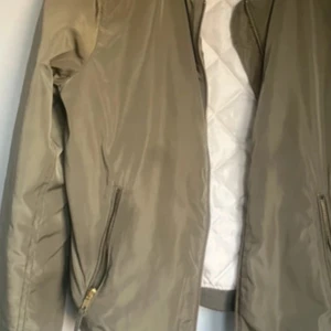 Olivgrön bomberjacka med dragkedjor - Snygg olivgrön bomberjacka med quiltat vitt foder. Jackan har flera dragkedjor, bland annat på ärmen och vid fickorna. Klassisk bombermodell med ribbade muddar och en stilren look. Perfekt för dig som gillar streetstyle.