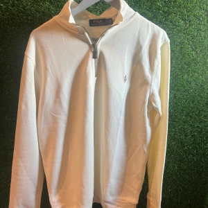 Vit half zip tröja från Polo Ralph Lauren - Snygg vit långärmad tröja från Polo Ralph Lauren med half zip och klassisk broderad logga på bröstet. Tröjan är i bomull och har en stilren, normal passform som funkar till många olika looks. Perfekt för dig som gillar clean och tidlös stil.