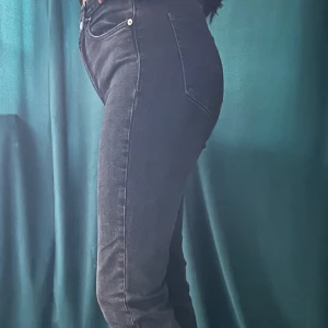 Mörkgrå bootcut jeans med slits - Säljer ett par mörkgrå jeans med bootcut passform och hög midja. Jeansen har klassiska fem fickor, dragkedja och knapp framtill samt snygga slitsar nertill vid bensluten. Perfekta för dig som gillar en tidlös men trendig look. (Priset går att diskutera)