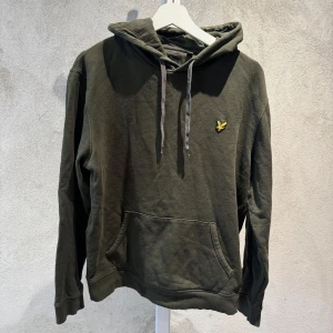 Mörkgrön hoodie från Lyle & Scott - Säljer en mörkgrön hoodie från Lyle & Scott i storlek L. Tröjan har huva med grå snörning, klassisk känguruficka och den ikoniska gula fågeln broderad på bröstet. Perfekt för chill dagar och enkel att matcha med jeans eller joggers.