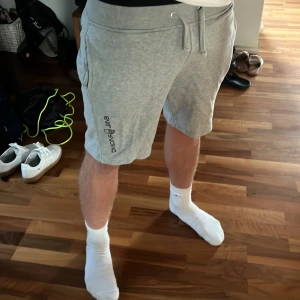 Grå Sail Racing shorts med snörning - Snygga grå shorts från Sail Racing med broderad logga på vänster ben. Modellen har midwaist passform, mjuk bomullskvalitet och praktisk snörning i midjan. Fickor på sidorna och en bakficka ger extra funktion. Perfekta för chill eller träning under sommaren.