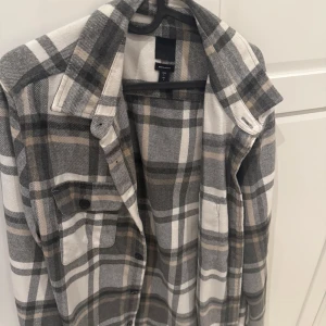 Rutig overshirt från H&M - Snygg rutig overshirt från HM i grått, vitt och beige. Skjortan har klassisk krage, bröstficka med knapp och knäppning framtill. Perfekt lager-på-lager-plagg med lite loose fit och mjukt bomullsmaterial.