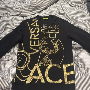Svart sweatshirt Versace Jeans guldtryck - Svart långärmad sweatshirt från Versace Jeans med stort guldtryck framtill, inklusive lejonmotiv och logga. Tröjan har rund halsringning och ribbade muddar. Perfekt för dig som vill sticka ut med en lyxig streetstyle.