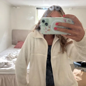 Vit teddyjacka - Mysig vit teddyjacka i fluffigt material med hel dragkedja framtill och hög krage. Så mysig flis till hösten. Jackan har lång ärm och två sidofickor, perfekt för lager-på-lager. Enkel och stilren design som passar till många outfits.