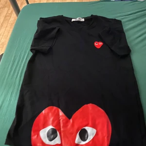 Svart Comme des Garçons Play t-shirt - Svart t-shirt från Comme des Garçons Play med klassiskt rött hjärta med ögon på bröstet och stort tryck nertill. T-shirten är i mjuk bomull och har en normal passform med korta ärmar. Perfekt för dig som gillar streetstyle och ikoniska prints.