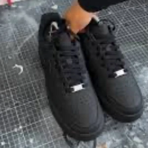 Svarta Nike Air Force 1 sneakers - Snygga helsvarta Nike Air Force 1 sneakers med klassisk siluett och perforerad tå. Skorna har svart snörning och en platt sula, perfekta för en clean streetstyle-look.