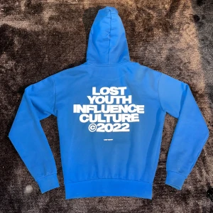 Blå hoodie Lost Youth Influence Culture - Storlek XS men lite oversized passform, hård/tajt resor i midjan vilket gör att man kan lägga tröjan vart man vill✅Knappast använd och har nog de bästa materialet och passform på en tröjan jag nånsin haft.✅                                  Nypris: 1300kr✅ Säljer en blå hoodie från Lost Youth med stor vit text 'Lost Youth Influence Culture ©2022' på ryggen och mindre tryck på bröstet. Tröjan har huva, känguruficka och långa ärmar. Perfekt för dig som gillar streetwear och bold prints.