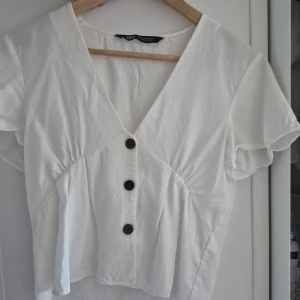 Vit blus med v-ringning från Zara - Säljer en vit blus från Zara med snygg v-ringning och tre svarta knappar framtill. Blusen har korta ärmar 🤍🎀