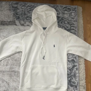 Vit hoodie från Polo Ralph Lauren - Snygg vit hoodie från Polo Ralph Lauren med klassisk broderad logga på bröstet. Tröjan har huva med snörning och en stor magficka. Perfekt för dig som gillar stilren och enkel design med premiumkänsla.