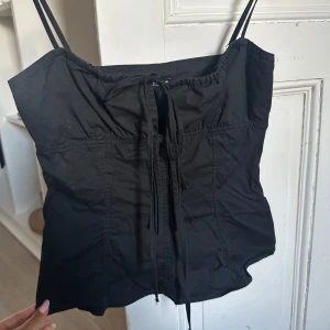 Svart linnetopp med snörning från Zara - Svart linnetopp från Zara med tunna axelband och snygg snörning framtill. Toppen har en rak passform och är tillverkad i bomull, perfekt för en chill och stilren look. Enkel att matcha med både jeans och kjol.