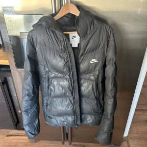 Svart pufferjacka från Nike - Svart pufferjacka från Nike med huva och två sidofickor med dragkedja. Jackan har ett stilrent, sportigt utseende och är vadderad för extra värme. Nike-loggan är broderad på bröstet. Perfekt för kalla dagar och enkel att matcha med streetwear. Några små hål på ena ärmen och framsidan men enkelt att laga