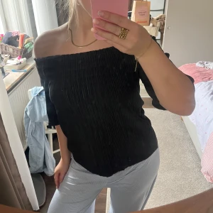 Svart offshoulder topp med plissering - Snygg svart offshoulder topp med plisserad struktur och trekvartsärm. Toppens elastiska kant gör att den sitter på plats över axlarna och ger en trendig look. Perfekt att styla med både jeans och kjol för en cool vibe.