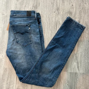 Replay Anbass blå jeans - Snygga Replay Anbass jeans i klassisk blå tvätt med diskret slitning. Modellen har raka ben, fem fickor och tydliga Replay-detaljer på fickorna. Jeansen är gjorda i mjukt denimtyg med stretch för extra komfort. 30/32
