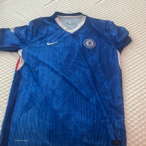 Chelsea FC Palmer 20 matchtröja Nike - Chelsea FC hemmatröja med Palmer 20-tryck, tillverkad av Nike i blått funktionsmaterial. Tröjan har korta ärmar, V-ringning och diskret randigt mönster. Premier League-märke på ärmen och klubbmärke på bröstet. Perfekt för dig som älskar fotboll och Chelsea.