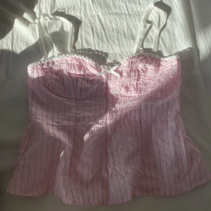 Rosa randig korsett-topp med rosett - Supersöt rosa korsett-topp med tunna vita och rosa ränder, vita axelband och liten rosett framtill. 