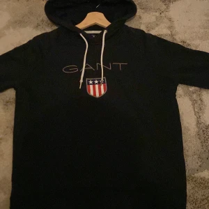 Svart hoodie från GANT med logga - Svart hoodie från GANT med broderad logga och sköld på bröstet. Tröjan har vit snörning i huvan och klassisk passform. Perfekt för dig som gillar stilren streetwear med en sportig touch, tröjan är i nytt skick.