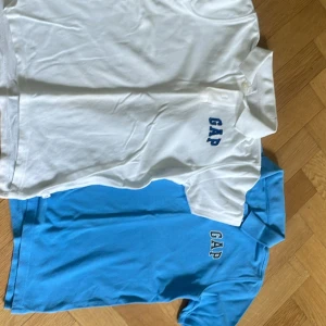 2-pack GAP pikétröjor vit & blå - Två klassiska pikétröjor från GAP, en i vitt och en i klarblått. Båda har kort ärm, krage och knäppning framtill samt GAP-logga broderad på bröstet. Perfekt för en clean och sportig look. Materialet är mjuk bomull som andas.