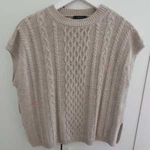 Beige stickad väst från Trendyol - Beige stickad väst med kabelstickade detaljer och rund hals. Oversized passform med slitsar i sidorna för en chill vibe. Perfekt att slänga över en skjorta eller t-shirt för en snygg lager-på-lager-look. Kommer med bälte (bältet syns ej på bild men tillhör tröjan).