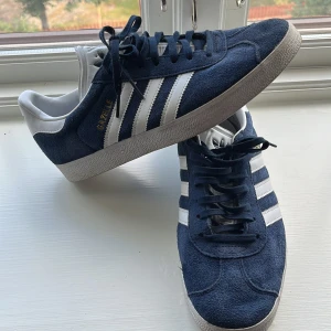 Adidas Gazelle blå/vita sneakers - Snygga Adidas Gazelle sneakers i mörkblå mocka med vita detaljer och klassiska tre ränder på sidorna. Skorna är använda en gång. Ny pris 1200 mitt pris 500 men inte hugget i sten👍👌