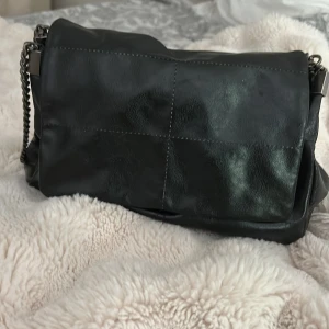 Svart axelväska i skinn med kedja från ZARA - Snygg svart axelväska i skinn med silverfärgad kedjerem och flera fack inuti. Väskan har stilrena sömmar som ger en cool känsla och dragkedja för extra säkerhet. Perfekt storlek för att få plats med allt du behöver. Älskar denna väska från Zara !