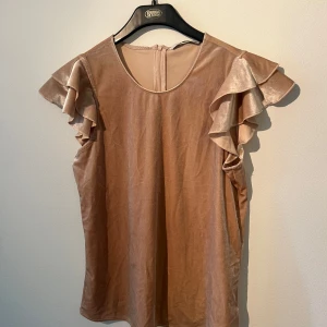 Beige sammetsblus med volangärm ONLY - Supersöt beige sammetsblus från ONLY i storlek 38. Blusen har korta volangärmar och rund halsringning. Materialet är mjukt och skimrande, vilket ger en lyxig känsla. Perfekt för dig som vill sticka ut med en feminin och trendig look.