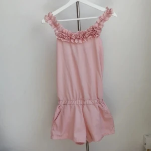 Rosa kort jumpsuit med volang - Supersöt ljusrosa kort jumpsuit med volangdetaljer runt axelbanden och halsringningen. Ärmlös och har en loose passform med resår i midjan och fickor. Passar xs-s, jag är i storlek S 160cm på bild 3. Använd enstaka gång.