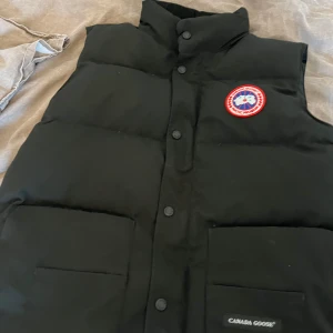 Svart dunväst från Canada Goose L - Svart dunväst från Canada Goose med klassisk logga på bröstet. Västen har hög krage, stora fickor framtill och stängs med knappar. Perfekt för lager-på-lager och riktigt snygg till streetwear-stil.