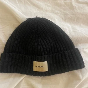 Svart ribbstickad mössa från Dondup - Svart ribbstickad mössa från Dondup med bred uppvikt kant och tydlig logotyp-patch framtill. Klassisk form som sitter snyggt på huvudet och passar perfekt till kalla dagar. Tillverkad i Italien och känns riktigt lyxig och stilren. One size.