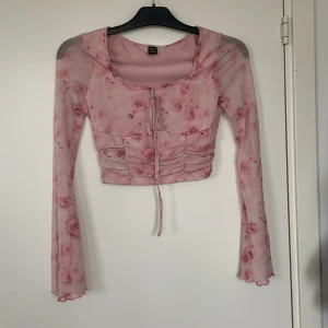 Rosa mesh topp med blommigt mönster - Supersöt croppad blus i ljusrosa med blommigt mönster och långa ärmar. Toppen har snörning framtill och lätt rynkade detaljer vid magen!