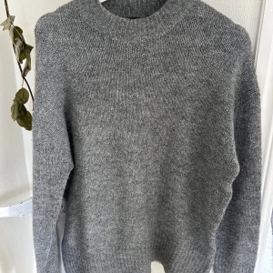 Grå stickad tröja med rund hals - Mysig grå stickad tröja med rund hals och långa ärmar. Perfekt för kyliga dagar och enkel att matcha med jeans eller kjol. Tröjan har en klassisk, lite loose passform och ribbade muddar vid hals, ärmslut och nederkant.
