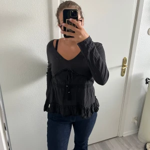 Svart blus med volang från Odd Molly - Supersöt svart blus från Odd Molly med långa ärmar, v-ringning och volangdetaljer längst ner. Blusen har snörning med tofsar och pärlor framtill samt spetsband över bröstet. Jätte skönt material