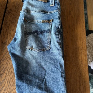 Blå jeans från Nudie Jeans - Snygga blå jeans från Nudie Jeans med klassisk femficksdesign och coola broderade detaljer på bakfickorna. Jeansen har en smal passform och är tillverkade i mjukt jeansmaterial med lätt tvättad look och slitningar på benen. Perfekt för dig som gillar stilrena och moderna jeans.skriv om du har funderingar 