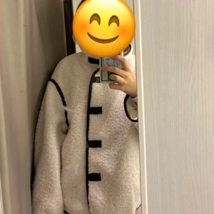 Vit teddyjacka med svarta detaljer - Supermysig vit teddyjacka med svarta kontrastdetaljer längs kanterna och knäppningen. Jackan har oversized passform och långa ärmar, perfekt för kalla dagar. Stängs med svarta knappar framtill och har en cool, unik look. Köpt från H&M förra året och den sålde slut snabbt!