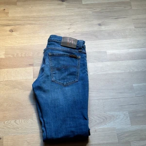Blå slim fit jeans från Nudie Jeans - Snygga blå jeans från Nudie Jeans med klassisk femficksdesign och orangea sömmar. Modellen har rak slim fit och normal midja, med ett brunt läderpatch med Nudie-loggan bak. Perfekta för dig som gillar stilrena och tidlösa jeans.skriv vid minst funderin