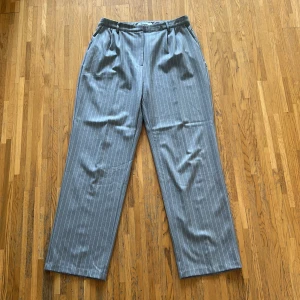 Grå randiga kostymbyxor från NA-KD - Snygga grå kostymbyxor från NA-KD med vita ränder. Byxorna har en klassisk rak passform och är perfekta för en stilren look. De har en hög midja och stängs med dragkedja och knapp.