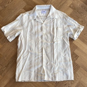 Randig kortärmad skjorta från NN07 - Säljer en stilren kortärmad skjorta från NN07 i en ljus beige och vit randig design. Skjortan har en klassisk krage och knappar framtill. Perfekt för en avslappnad och somrig look.