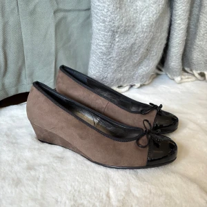 Bruna kilklackskor från Jenny - Snygga bruna kilklackskor från Jenny med svart lackad tå och rosett. Perfekta för en stilren look med en bekväm kilklack. Skorna har en elegant design med en kombination av mocka och lackdetaljer.