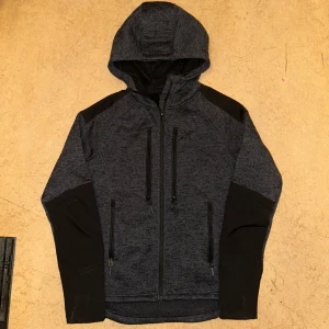 Mörkgrå hoodie från RevolutionRace - Säljer en mörkgrå hoodie från RevolutionRace med huva och dragkedja. hoodien har flera praktiska fickor med dragkedjor och svarta detaljer på ärmarna. Perfekt för utomhusaktiviteter.